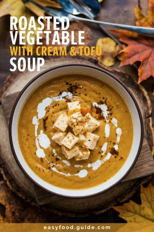 A bowl of creamy roasted vegetable soup with tofu, surrounded by autumn leaves on a wooden board.