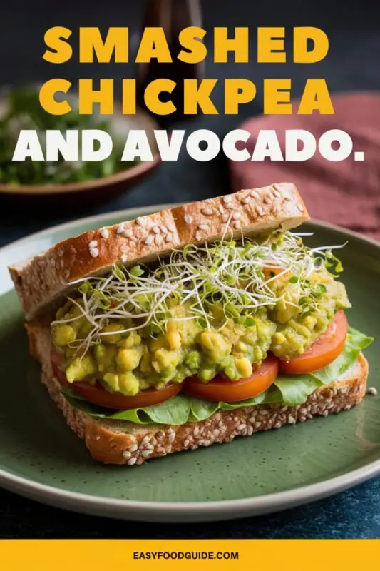 Smashed chickpea and avocado sandwich on a green plate with sprouts and tomato.