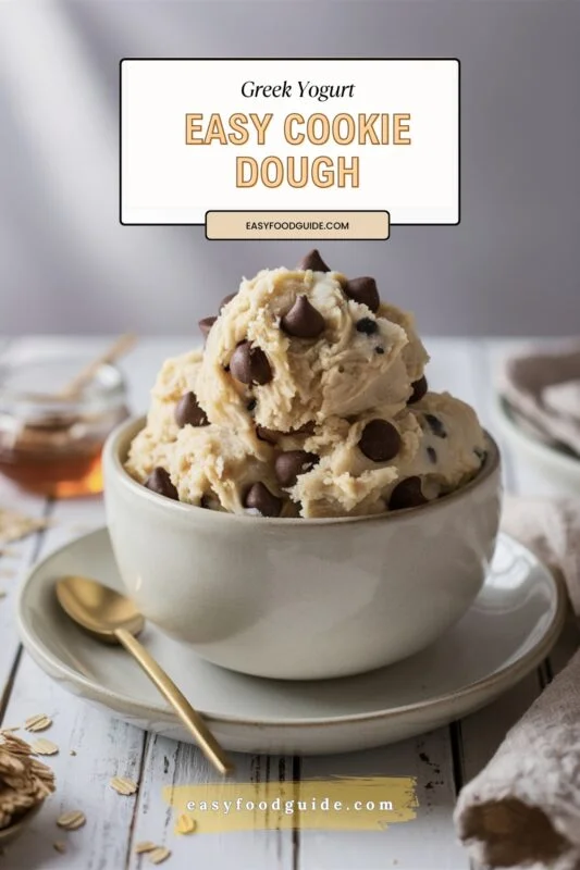 A bowl of creamy Greek yogurt cookie dough with chocolate chips styled elegantly on a rustic wooden surface, accompanied by a gold spoon and subtle background elements.