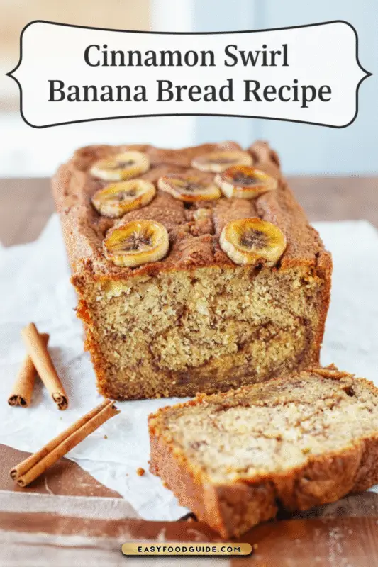 A loaf of cinnamon swirl banana bread with a slice cut to show the cinnamon sugar swirls, on a marble surface with cinnamon sticks and ripe bananas.