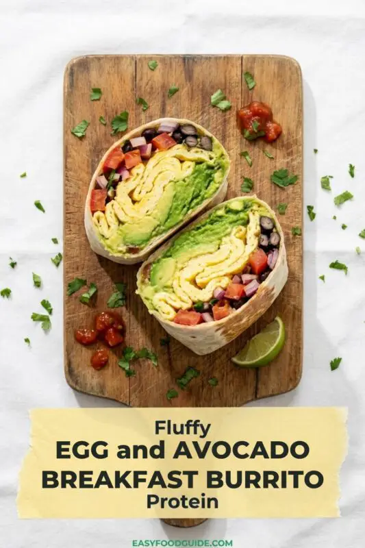 Overhead view of a halved egg and avocado breakfast burrito on a wooden board, packed with fluffy scrambled eggs, thick slices of avocado, black beans, and red peppers. The wrap is garnished with fresh cilantro and a lime wedge. Text overlay reads 'Fluffy EGG and AVOCADO BREAKFAST BURRITO Protein