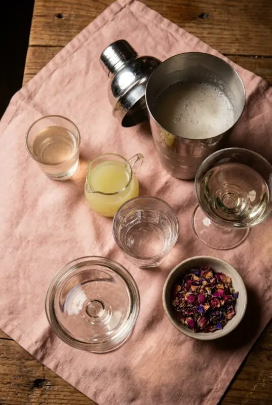 lychee-vodka-martini-4 Overhead view of lychee martini ingredients on a pink cloth, including a frothy metal cocktail shaker, coupe glasses, small glasses of clear and pale yellow liquid, a bowl of dried rose petals, and water on a rustic wooden table.