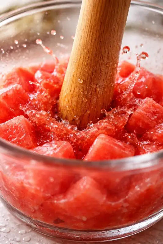 watermelon-tequila-smash-2b A wooden pestle crushes juicy watermelon cubes in a glass bowl, creating splashes of red juice. The scene conveys freshness and vibrancy.