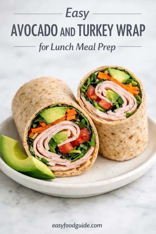 An image of an easy avocado and turkey wrap, cut in half to show the filling of rolled turkey, avocado, spinach, and carrots on a white plate with an avocado wedge. Text overlay reads "Easy AVOCADO AND TURKEY WRAP for Lunch Meal Prep," with the website "easyfoodguide.com" at the bottom.