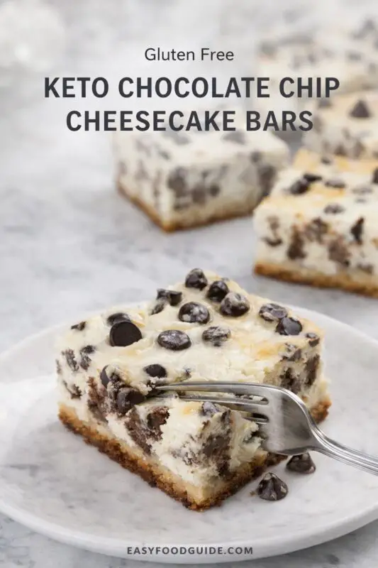 A square keto chocolate chip cheesecake bar on a plate with a fork holding a bite. It features a crumbly base, creamy texture, and chocolate chips.