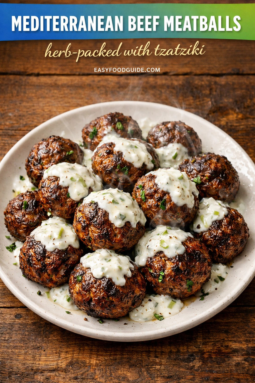 A rustic photograph of a plate of herb-packed, seared Mediterranean beef meatballs. The meatballs are generously topped with a creamy tzatziki sauce containing chopped green herbs. Piled on a speckled ceramic plate over a dark wooden surface. Top text banner on a blue-to-green gradient reads: 'MEDITERRANEAN BEEF MEATBALLS herb-packed with tzatziki' and 'EASYFOODGUIDE.COM'. Subtle steam rises.