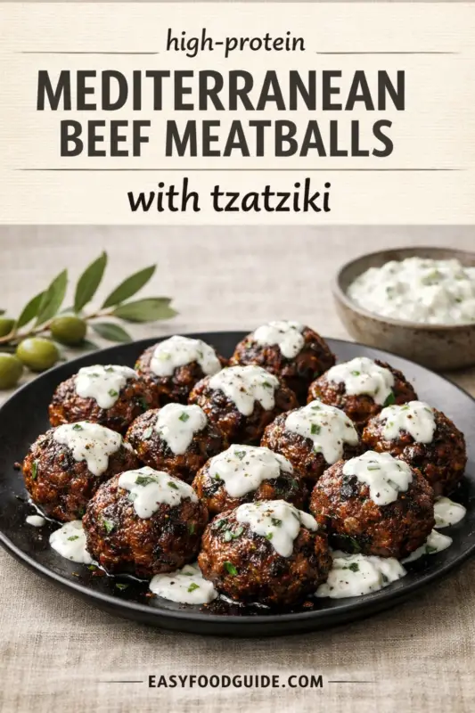 A plate of Mediterranean beef meatballs, topped with tzatziki and herbs. The title text reads 'high-protein MEDITERRANEAN BEEF MEATBALLS with tzatziki'. The scene includes green olives and an extra bowl of tzatziki. The URL 'EASYFOODGUIDE.COM' is at the bottom.