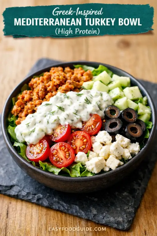 A top-down photograph of a dark ceramic bowl filled with a Greek-inspired Mediterranean turkey bowl. Arranged on a bed of lettuce are seasoned ground turkey, a creamy tzatziki sauce with dill, halved cherry tomatoes, cubed cucumbers, sliced black olives, and cubed feta cheese. The bowl rests on a dark slate slab on a wooden table. Overlaid text at the top reads, "GREEK-INSPIRED MEDITERRANEAN TURKEY BOWL (High Protein)" and at the bottom, "EASYFOODGUIDE.COM".