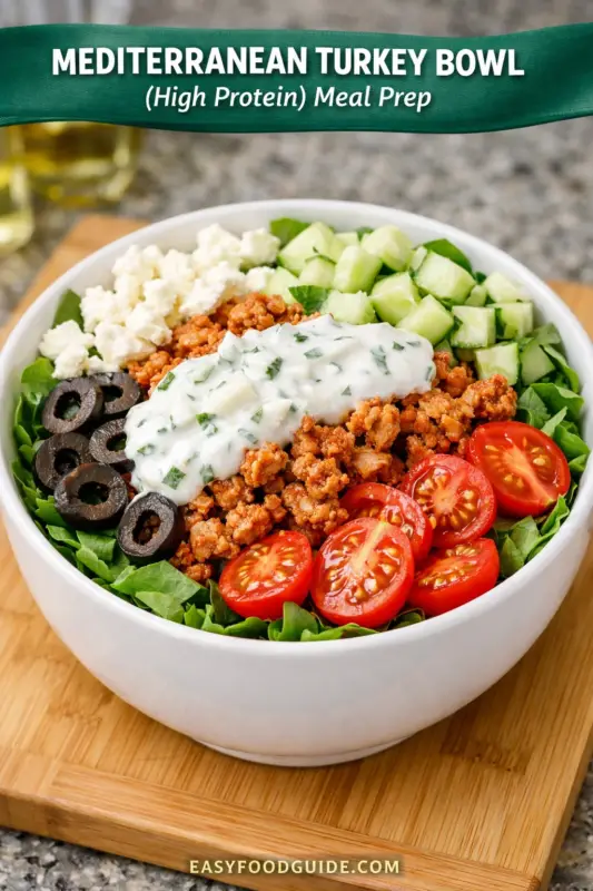 A photograph of a Mediterranean Turkey Bowl for meal prep, presented in a large white bowl on a wooden cutting board. The bowl is filled with layers of seasoned ground turkey, fresh spinach, cherry tomato halves, diced cucumbers, black olive slices, and crumbled feta cheese, all topped with creamy tzatziki sauce. A green banner across the top has text that reads "MEDITERRANEAN TURKEY BOWL (High Protein) Meal Prep". The text "EASYFOODGUIDE.COM" is at the bottom.