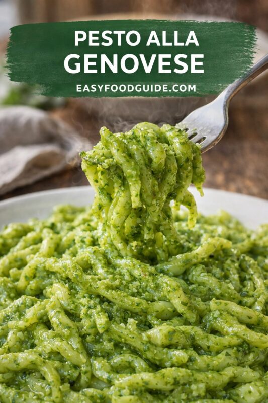 This image from EASYFOODGUIDE.COM features “PESTO ALLA GENOVESE.” A fork lifts steaming, vibrant green pesto-coated pasta — likely trofie or linguine — from a white bowl. The sauce is thick with visible basil, garlic, and nuts. Soft-focus background includes linen napkin and warm wood tones. Text overlay in bold white on brushed green banner highlights the classic Ligurian dish.